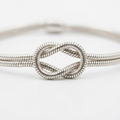 Silver-toned double snake chain bracelet infinity knot detail – two parallel cylindrical snake chains forming decorative love knot with hollow eye-shaped opening – extreme close-up product photography